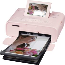 Ein rosa Canon SELPHY CP1300 Fotodrucker, bei dem das Druckfach ausgefahren und ein Foto einer lächelnden Frau, die Blumen hält, teilweise ausgedruckt zu sehen ist. Über dem Druckfach ist ein in Betrieb befindlicher Klappbildschirm zu erkennen, der ein digitalisiertes Spiegelbild derselben Frau zeigt, um zu demonstrieren, wie das gedruckte Bild ursprünglich auf dem Bildschirm aussah.