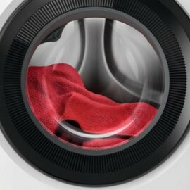 Das Bild zeigt eine Nahaufnahme der Frontansicht der AEG Lavamat LR7G60487 Waschmaschine in Weiß während des Betriebs. Im Fokus steht die geöffnete, transparente Bullaugentür, durch die man eine rote Textilie im Innern der Trommel erkennen kann, die anscheinend gerade gewaschen wird. Diese Darstellung dient dazu, das Design der Waschmaschine und ihre Funktionalität während des Waschvorgangs zu veranschaulichen.